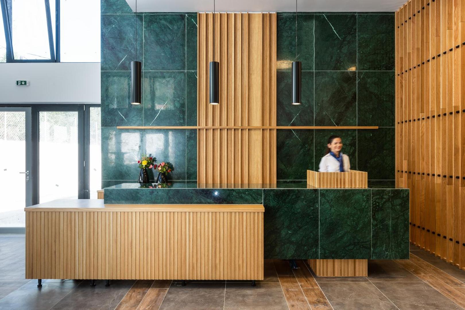 T Hotel Premium Suites recepcijas lete ar zaļā marmora un koka elementiem — viesnīcas darbiniece sagaida viesus