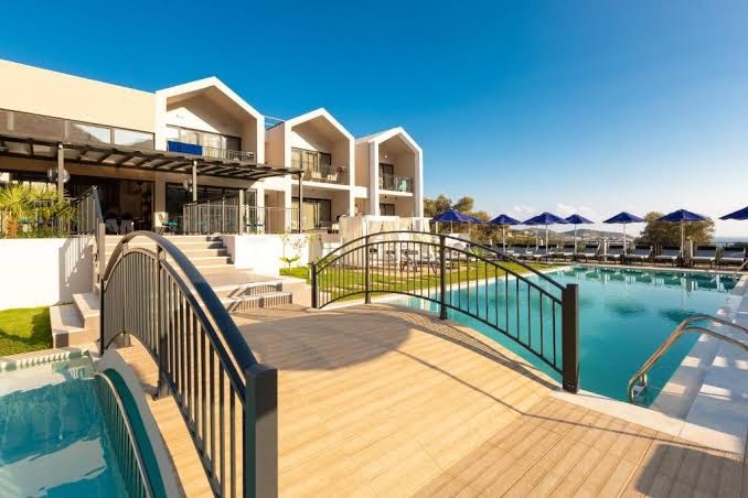T Hotel Premium Suites āra baseins ar koka tiltiņu un saulessargiem Bali ciematā, Krētā — skats uz modernu viesnīcas fasādi