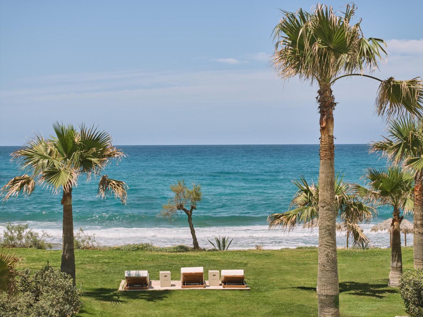 Skats no Caramel Grecotel Resort dārza uz zilo Vidusjūru Krētā ar palmām un atpūtas krēsliem zālienā pie pludmales
