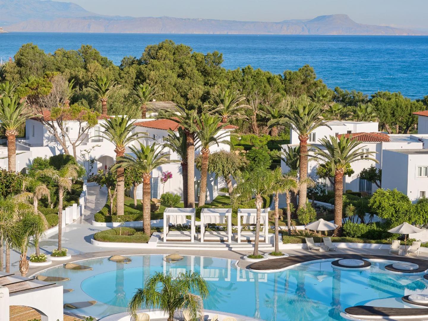 Caramel Grecotel Resort galvenais baseins ar atpūtas gultām un palmām Krētas piekrastē — fona skats uz jūru un kalniem