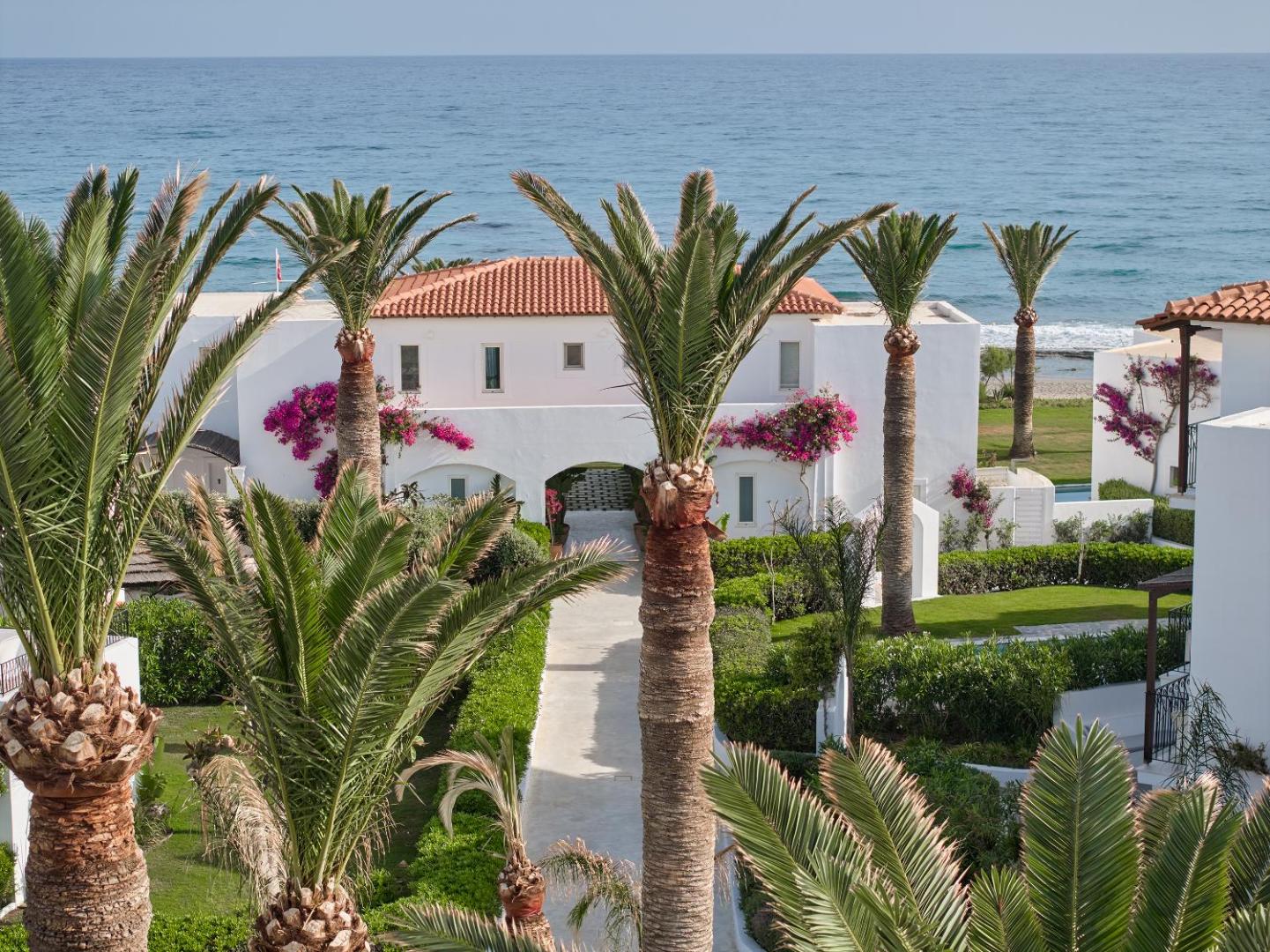 Gājēju celiņš starp palmām un ziedošiem krūmiem Caramel Grecotel Resort teritorijā Krētā ar skatu uz Vidusjūru