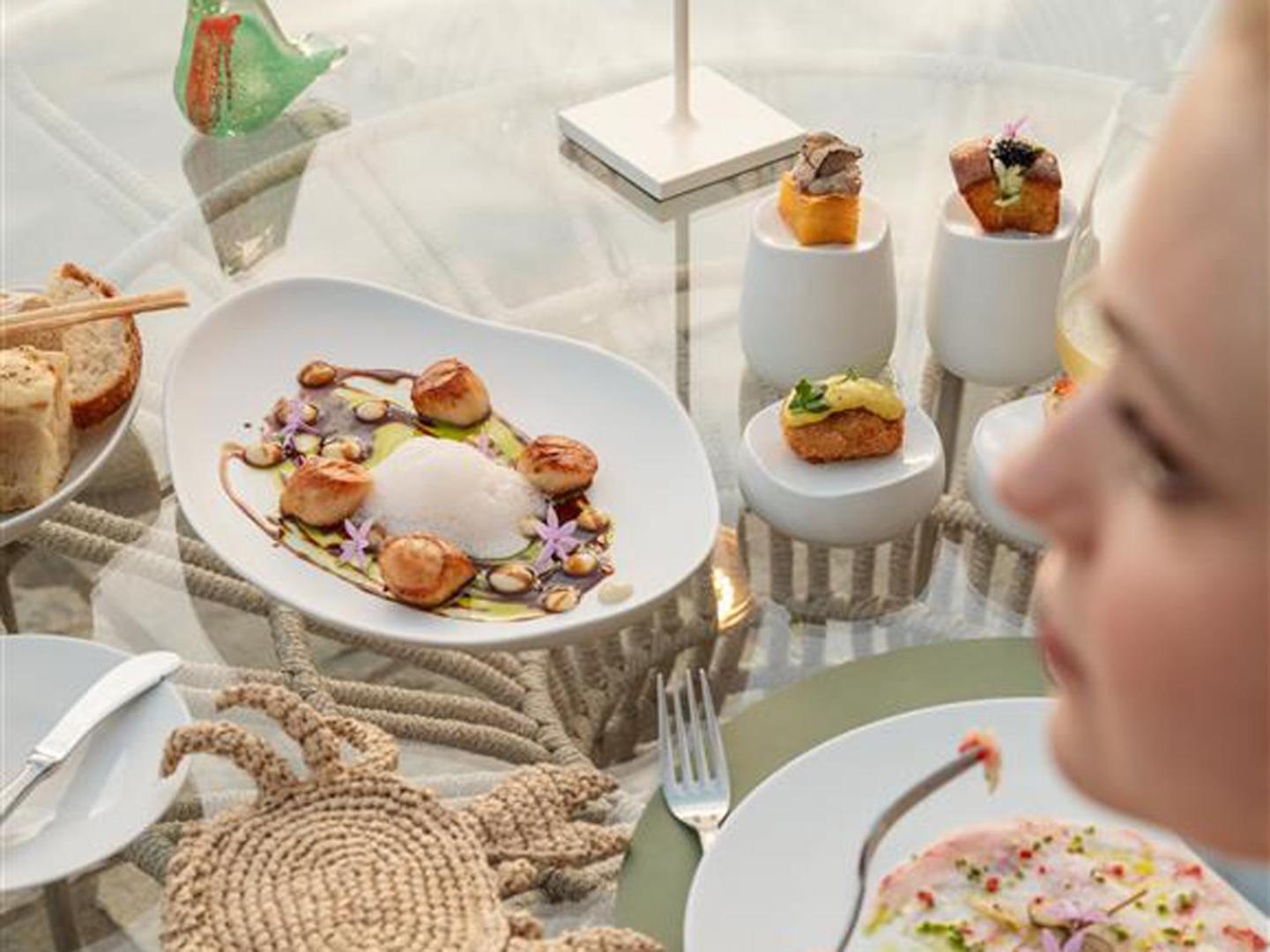 Izsmalcinātas uzkodas un jūras veltes uz stikla galda Caramel Grecotel Boutique Resort terasē