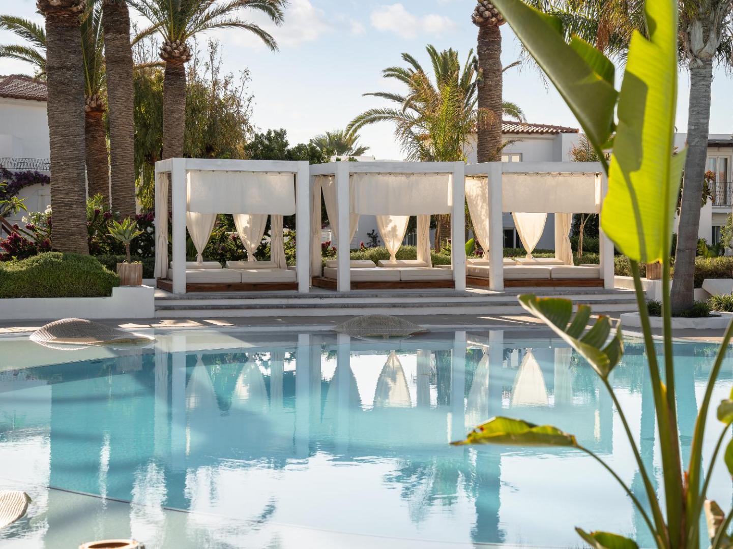 Grezni balti atpūtas bungalo pie Caramel Grecotel Resort baseina Krētā — mīksti matrači un aizkari privātumam palmu ielenkumā