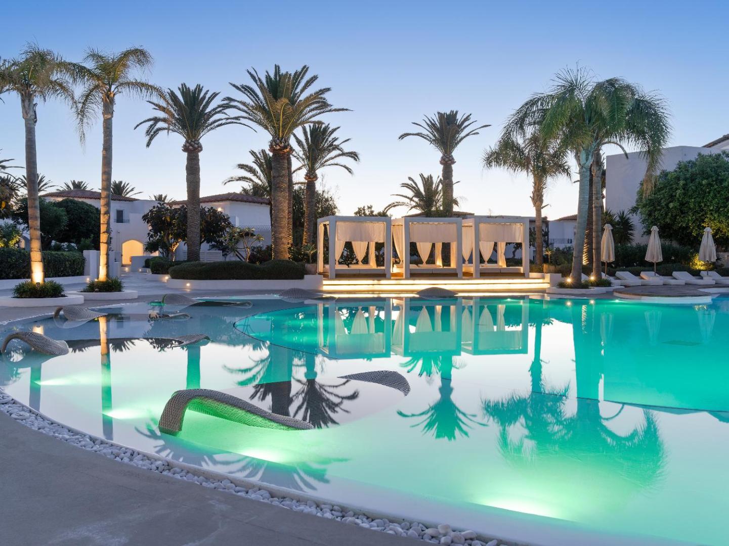 Caramel Grecotel Resort izgaismots āra baseins vakarā ar palmām un atpūtas nojumēm Retimno piekrastē, Krētā