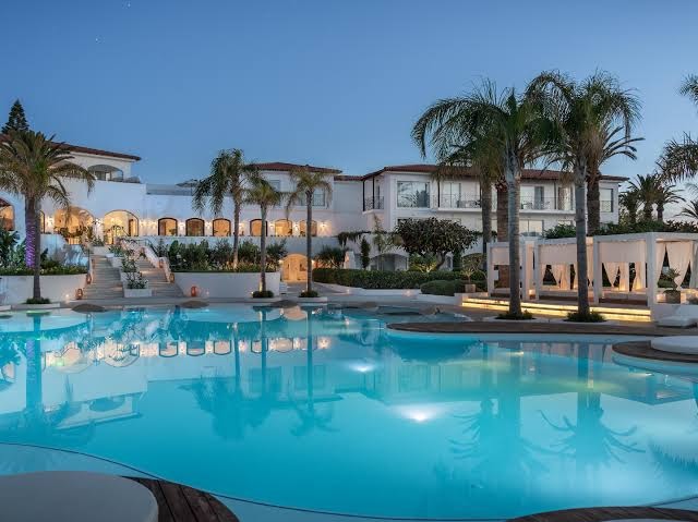 Caramel Grecotel Resort galvenā ēka un izgaismots baseins vakarā Retimno piekrastē — eleganta arhitektūra un palmas krēslas stundā