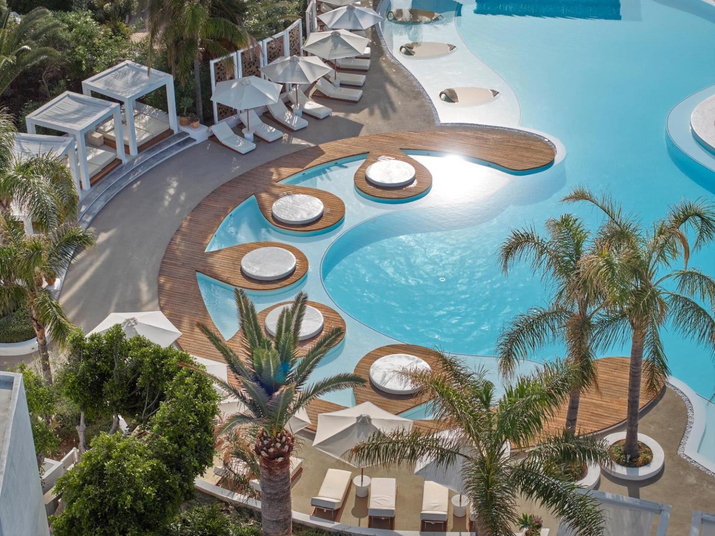 Skats no augšas uz Caramel Grecotel Resort baseina zonu — balti saulessargi, sauļošanās krēsli un koka detaļas pie tirkīza ūdens