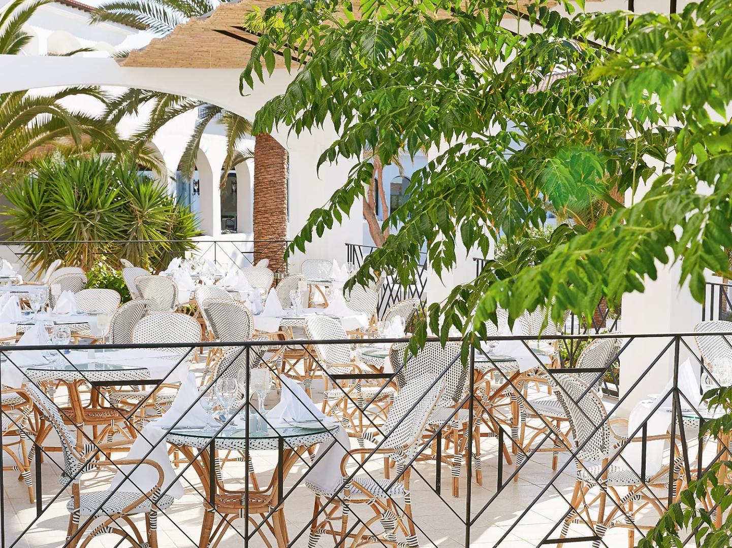 Gaiša viesnīcas Caramel Grecotel Resort restorāna terase ar pītiem krēsliem, baltiem galdautiem un zaļojošiem augiem saules gaismā
