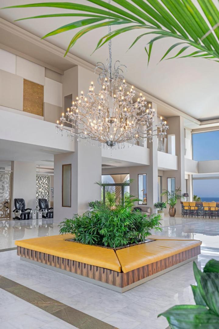 Kresten Royal Euphoria Resort vestibila atpūtas zona ar dzeltenu dīvānu, lielu kristāla lustru un eksotiskiem telpaugiem Rodā