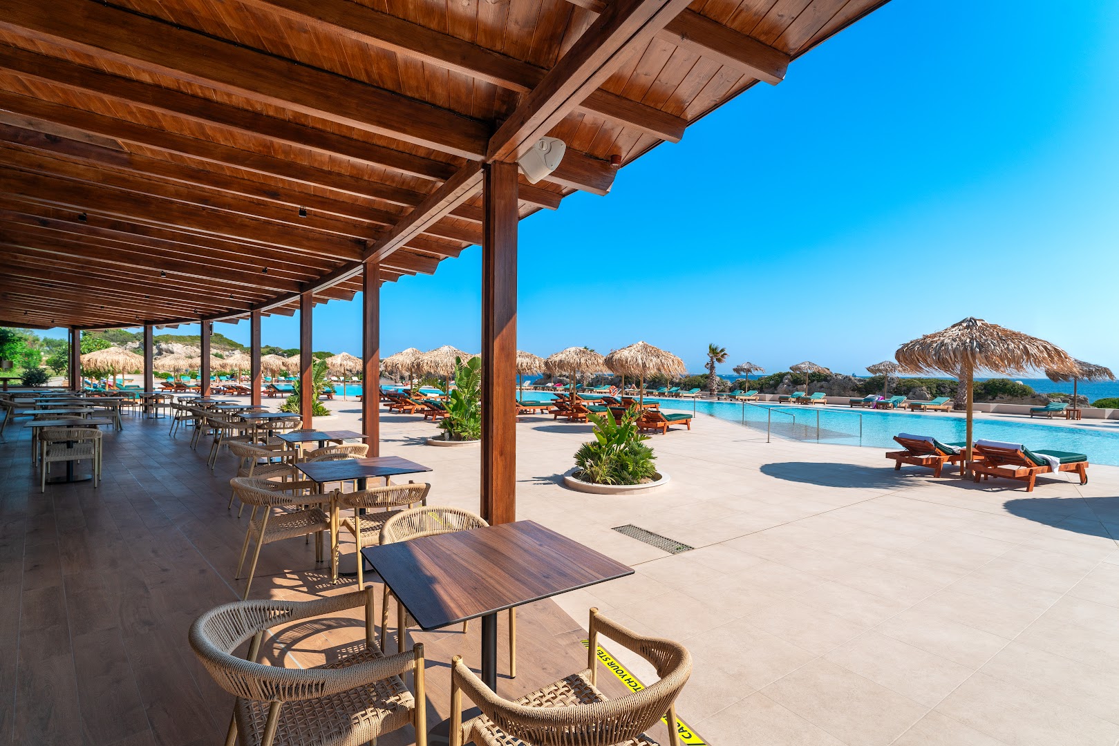 Kresten Royal Euphoria Resort āra terase ar galdiņiem pie baseina Rodā, Grieķijā — saulaina atpūtas zona ar salmu saulessargiem un zviļņiem