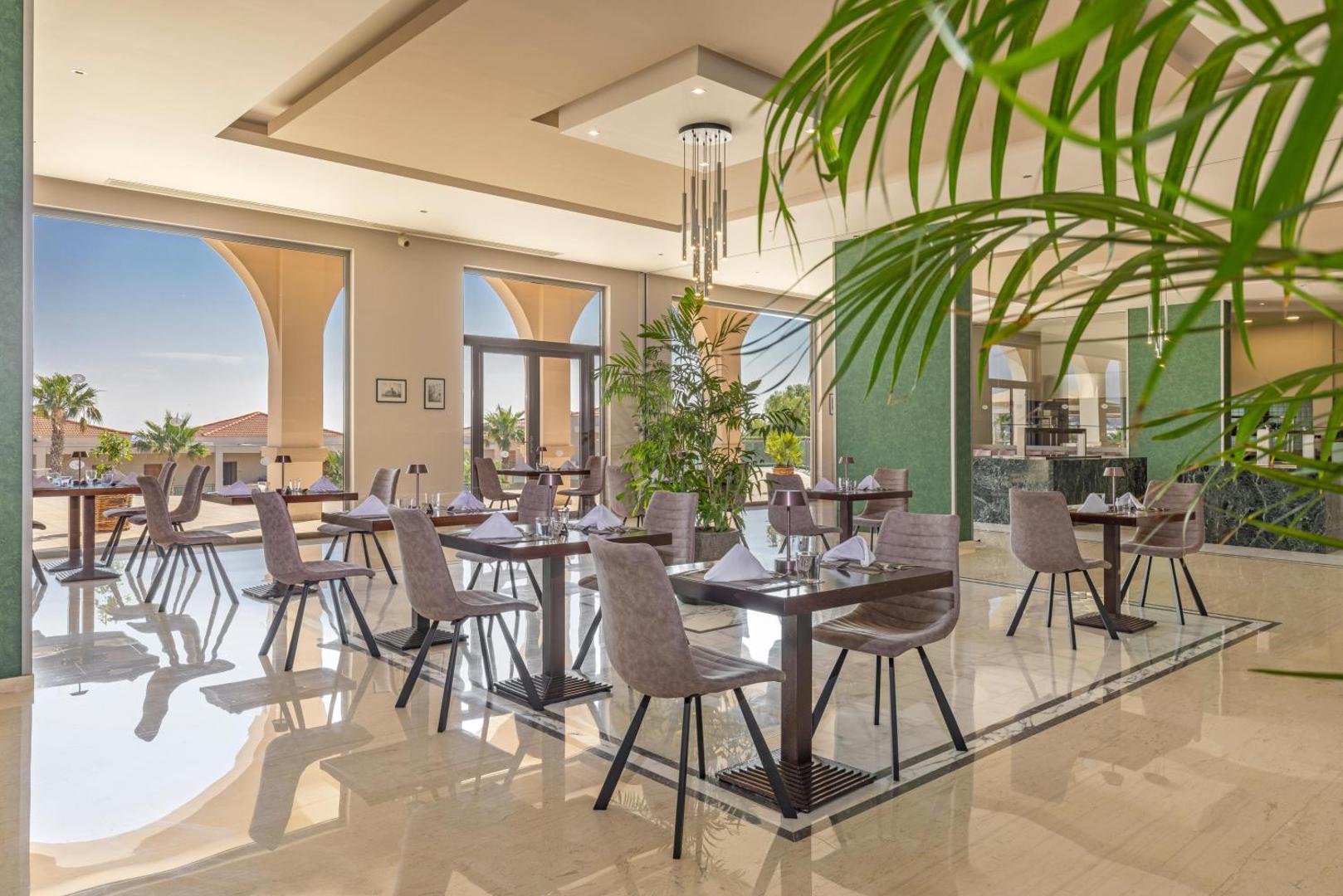 Kresten Royal Euphoria Resort elegantais ēdienkartes restorāns ar gaišu interjeru, lieliem logiem un skatu uz kūrorta teritoriju Rodā