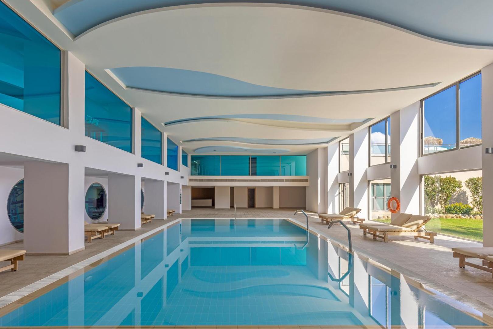 Kresten Royal Euphoria Resort iekštelpu baseins ar panorāmas logiem, gaišu dizainu un ērtiem zviļņiem relaksācijai Rodas salā