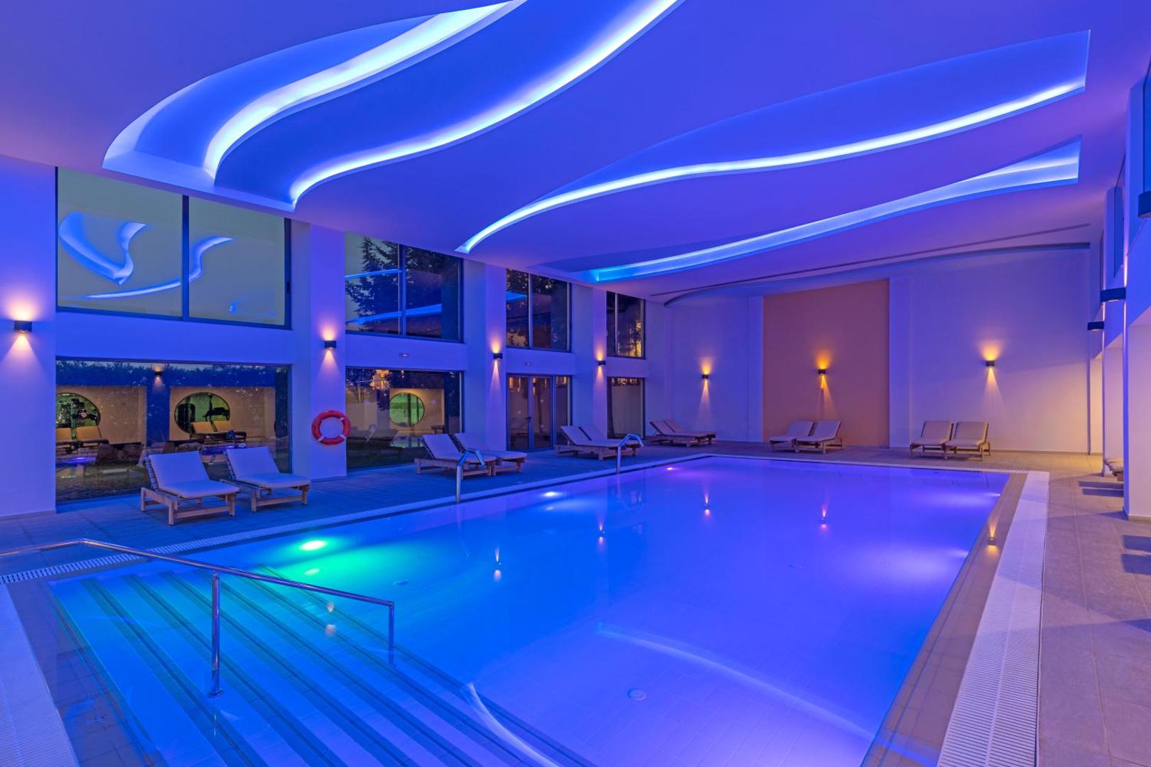 Kresten Royal Euphoria Resort spa zonas baseins vakarā ar zilu apgaismojumu un viļņveida griestu dizainu Rodā