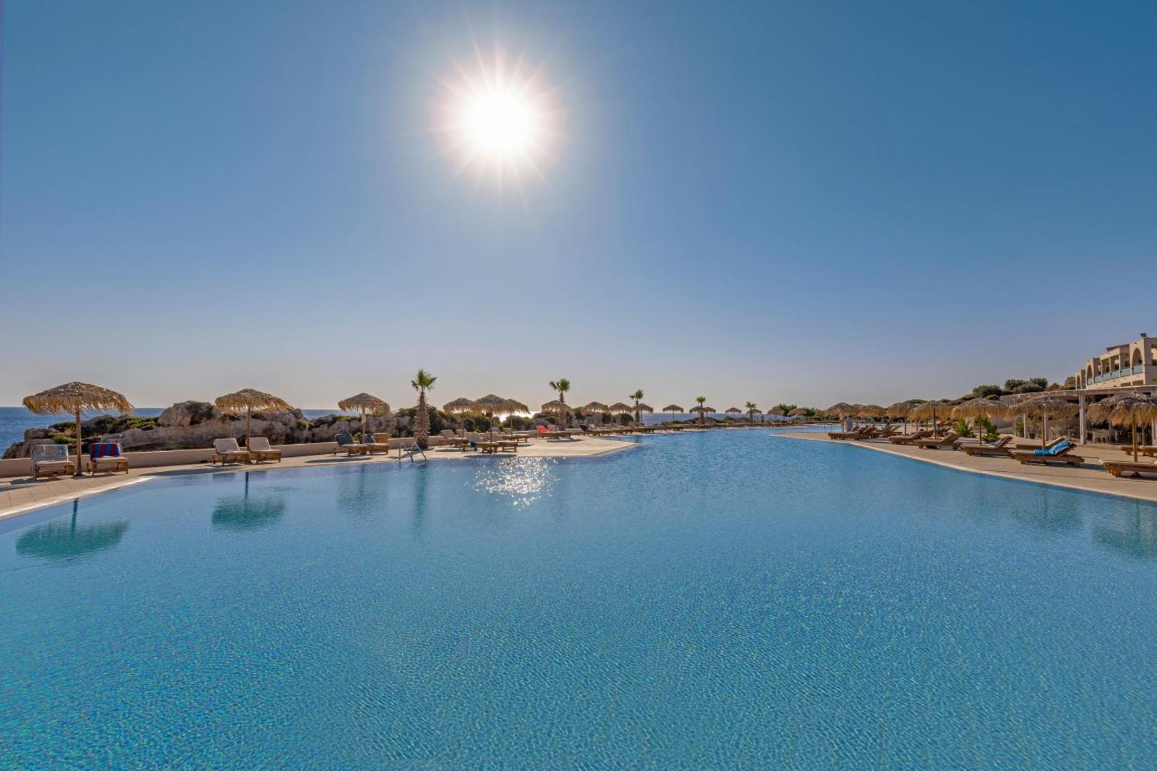 Kresten Royal Euphoria Resort plašais āra baseins saulainā dienā Rodā — piejūras ainava ar klintīm un salmu saulessargiem