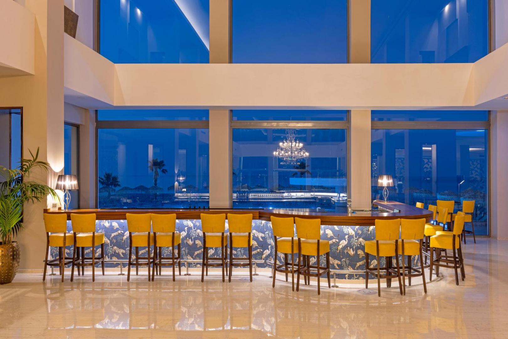 Kresten Royal Euphoria Resort lobby bārs ar dzelteniem krēsliem un lieliem logiem uz jūru vakara stundās Rodā