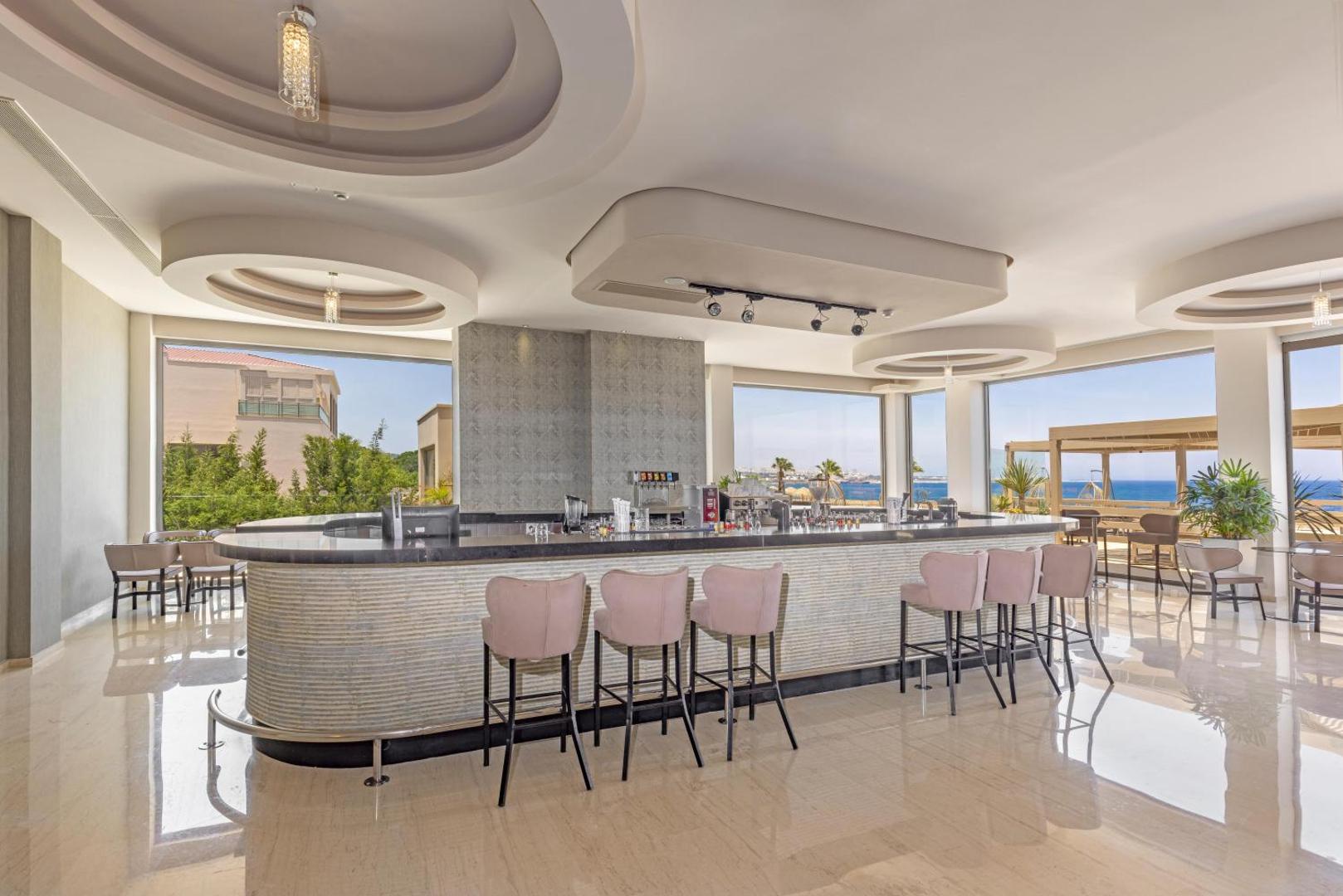 Kresten Royal Euphoria Resort modernais lobby bārs ar gaišiem krēsliem, plašiem logiem un skatu uz jūru Rodas salā, Grieķijā.