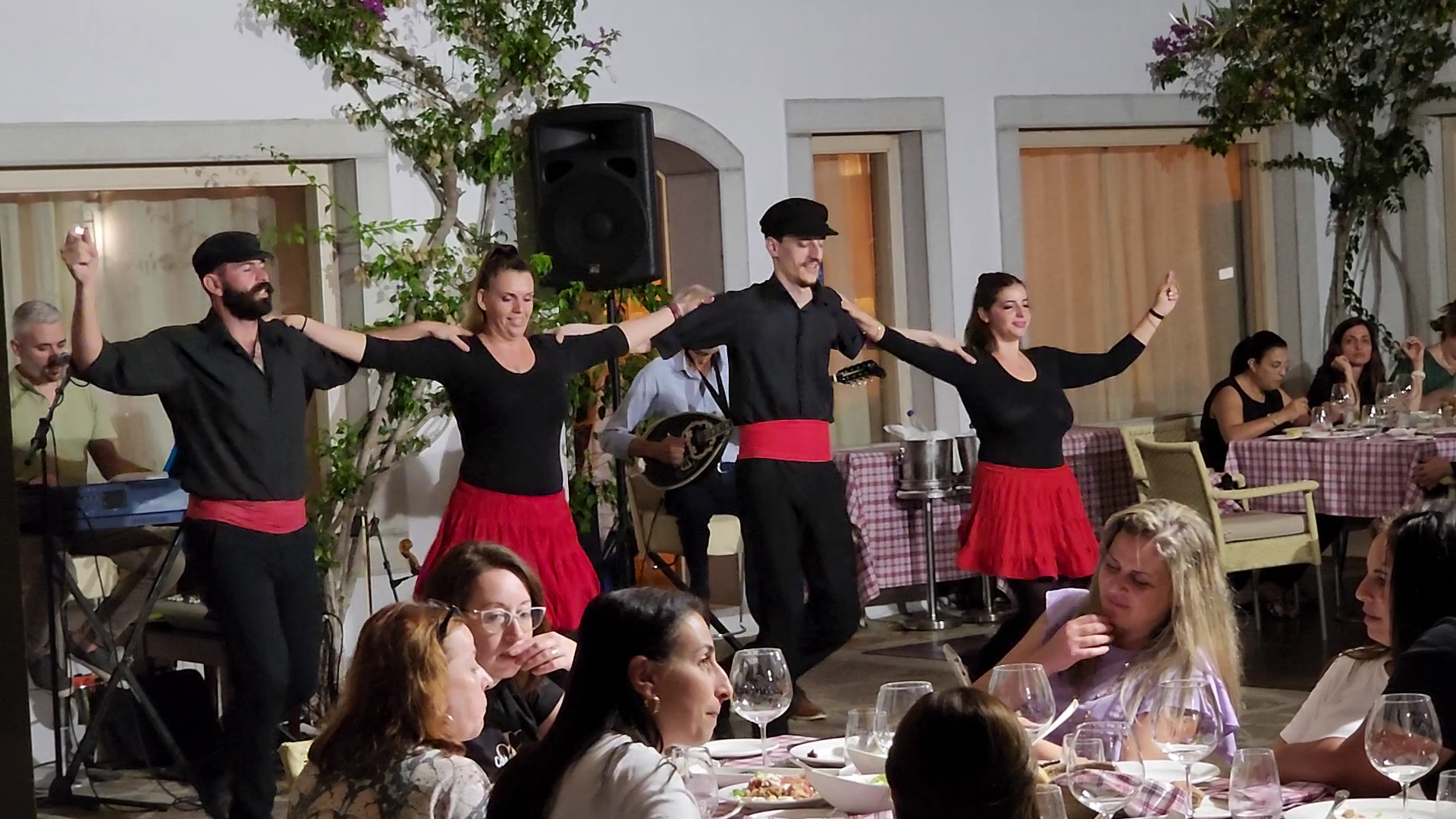 Grieķu tradicionālo deju priekšnesums Elounda Beach Hotel restorānā, dejotāji melnos tērpos ar sarkanām jostām izklaidē viesus