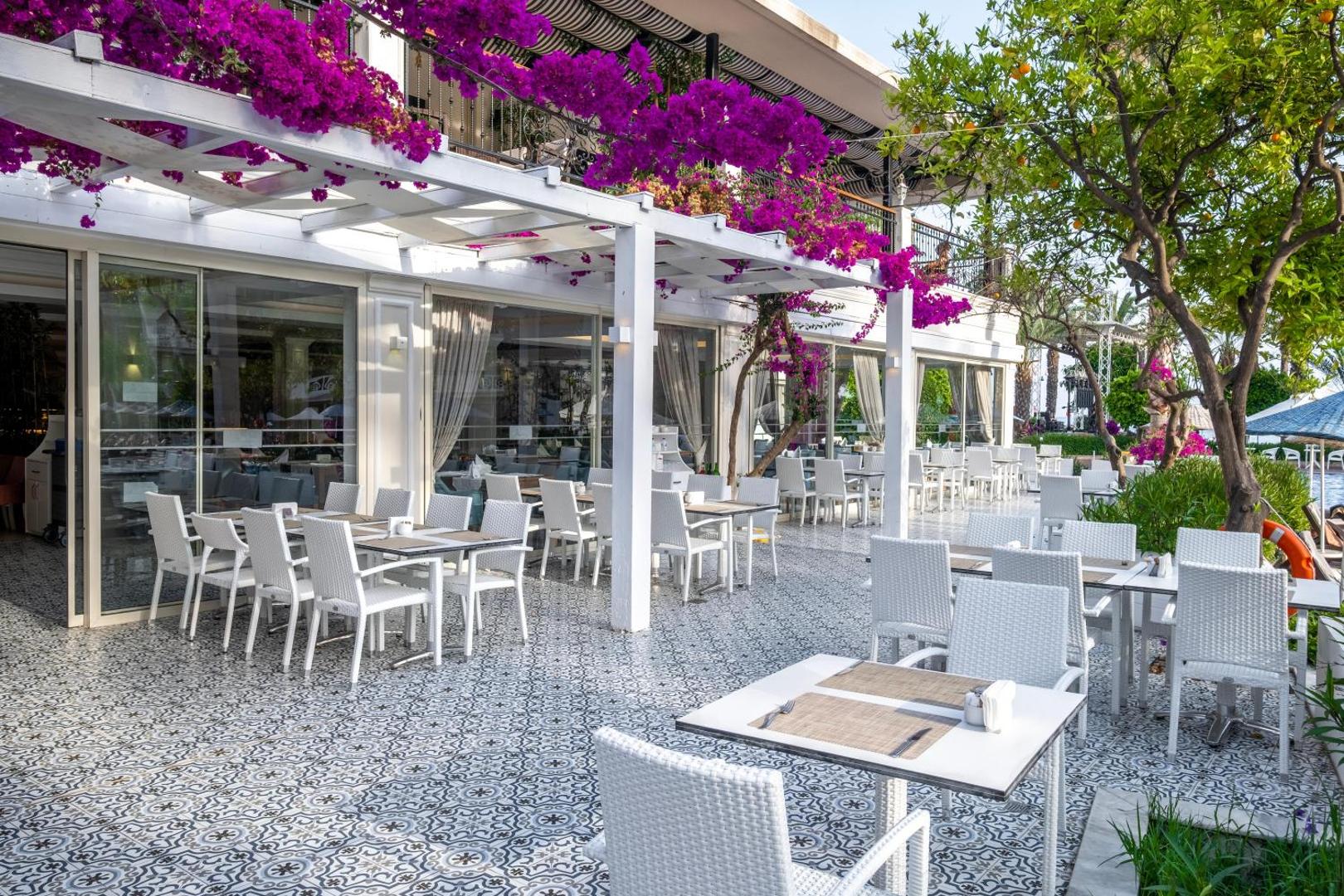 Utopia Resort & Residence āra terase ar baltiem pītiem krēsliem, rakstainām flīzēm un ziedošām bugenvillām piekrastē Turcijā.