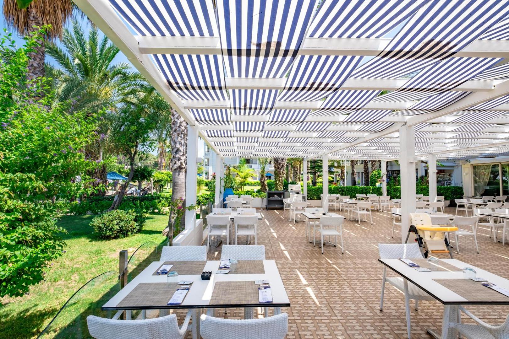 Utopia Resort & Residence vasaras restorāns zem svītrainas nojumes, ko ieskauj palmas un zaļš dārzs Alanjas piekrastē.