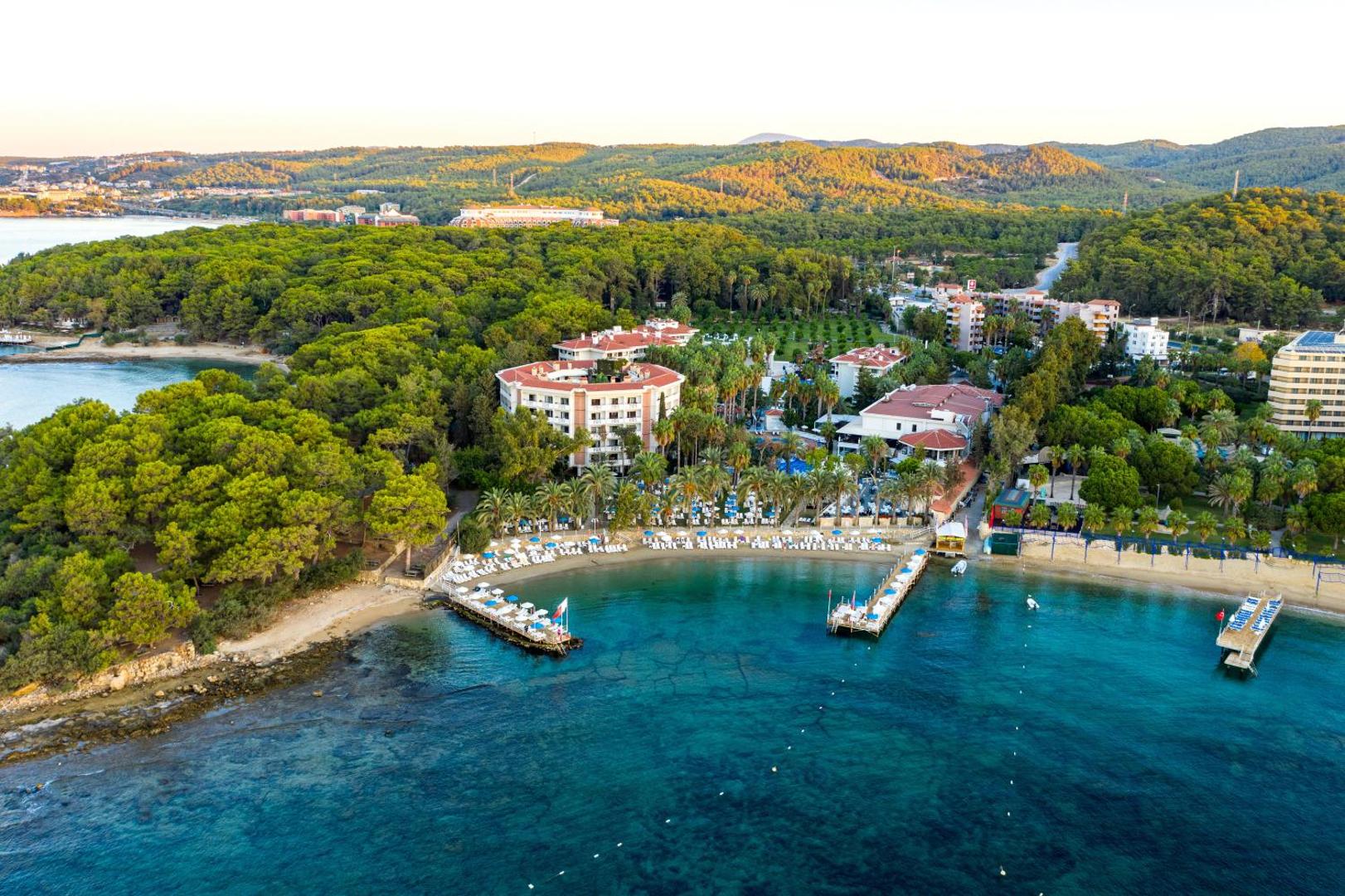 Utopia Resort & Residence panorāmas skats uz līci un viesnīcas ēkām, ko ieskauj blīvs priežu mežs un tirkīzzila jūra.