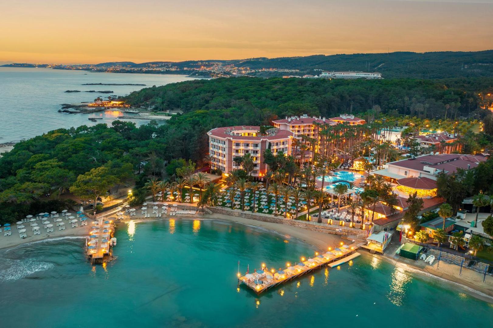 Utopia Resort & Residence skats no putna lidojuma saulrietā — izgaismota viesnīcas teritorija, baseini un koka piestātnes Alānijas piekrastē.