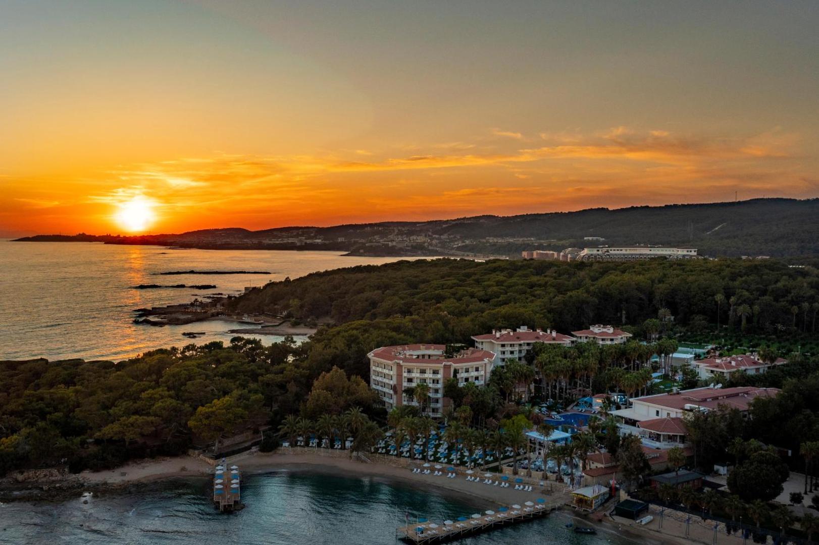 Utopia Resort & Residence skats no putna lidojuma saulrietā — viesnīcas korpusi, zaļojoša teritorija un privātais līcis Alānijas piekrastē
