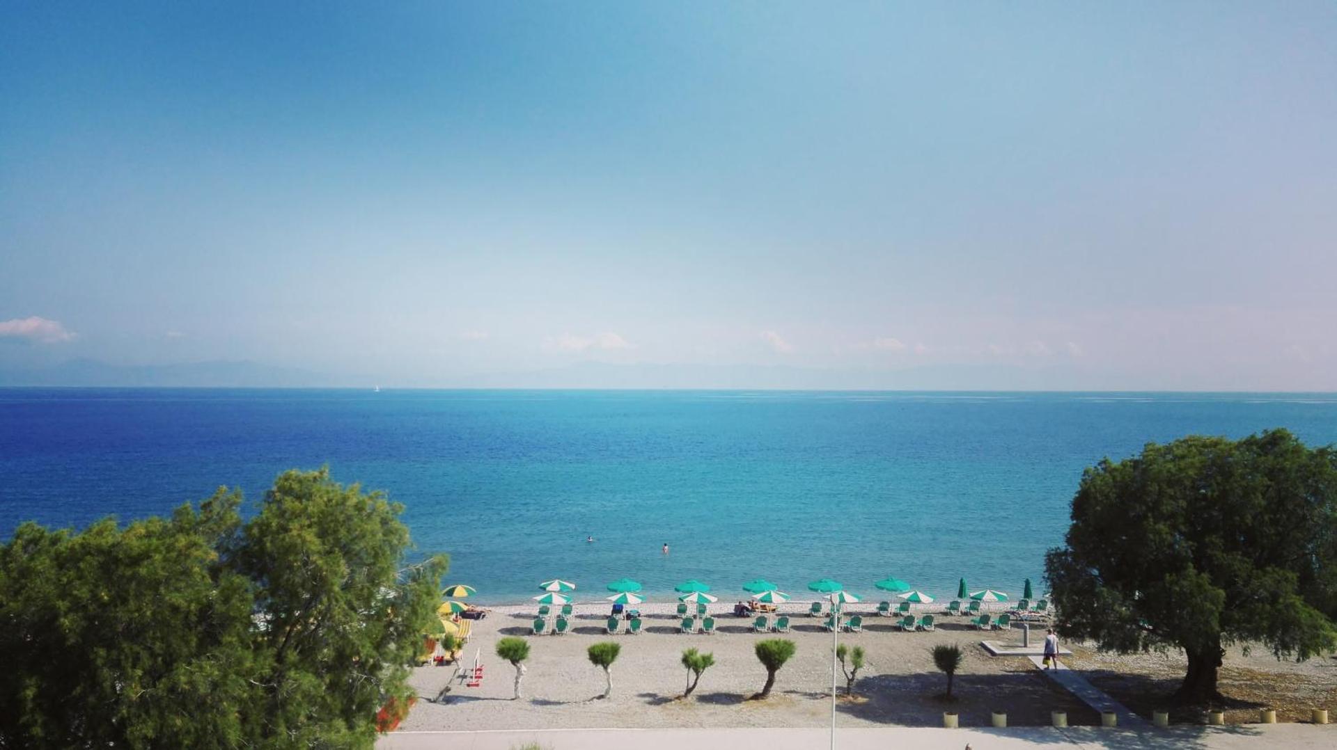 Pylea Beach Hotel panorāmas skats uz oļu pludmali ar saulessargiem un dzidri zilo Egejas jūru