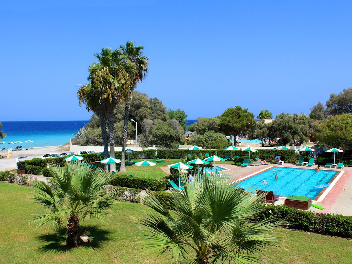 Pylea Beach Hotel teritorija ar palmām, koptu zālienu un baseinu, fonā Egejas jūras zilgme Rodas salas piekrastē.