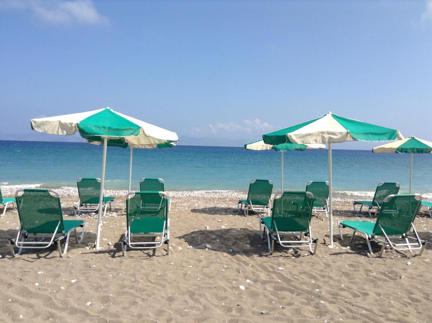 Pylea Beach Hotel plašā smilšu un oļu pludmale Rodā ar zaļiem sauļošanās krēsliem un saulessargiem pie dzidra ūdens