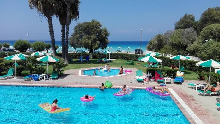 Viesi atpūšas Pylea Beach Hotel lielajā baseinā un bērnu baseinā, fonā redzamas palmas un zilā jūra piekrastē.