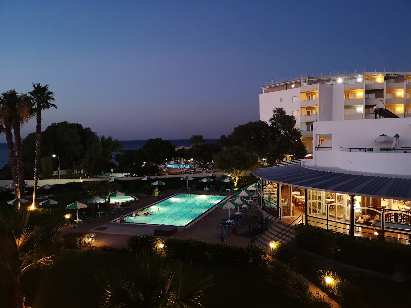 Pylea Beach Hotel vakara apgaismojums pie baseina un restorāna terases, skats uz izgaismoto viesnīcas ēku nakts laikā.