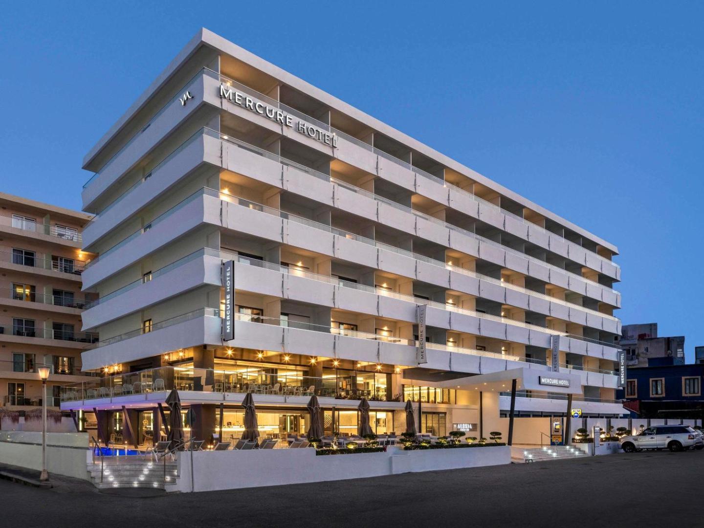 Mercure Rhodes Alexia Hotel & Spa skats no sāna vakarā — izgaismota terase, restorāna zona un baseina mala pilsētas vidē