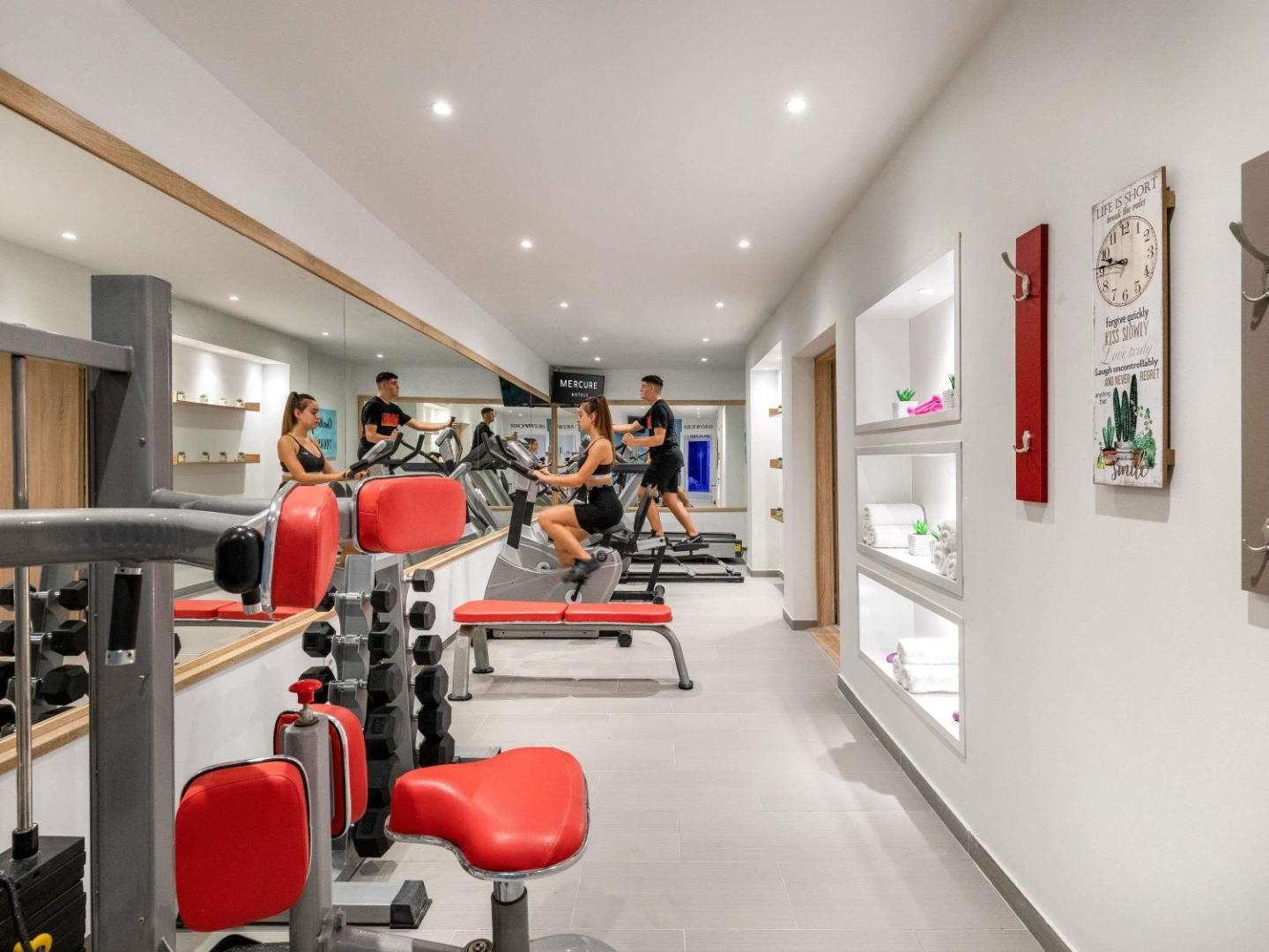 Mercure Rhodes Alexia Hotel & Spa fitness centrs ar moderniem trenažieriem, lielu spoguli un sarkanu akcentu aprīkojumu aktīvai atpūtai
