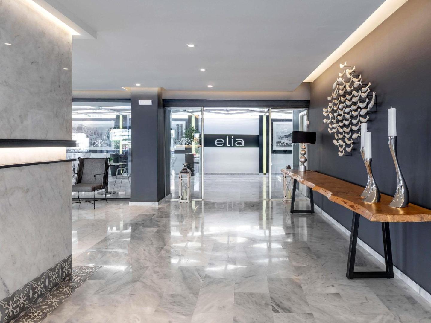 Ieeja Mercure Rhodes Alexia Hotel restorānā Elia ar stikla durvīm, marmora grīdu un masīvkoka dekoratīvo galdu pie sienas