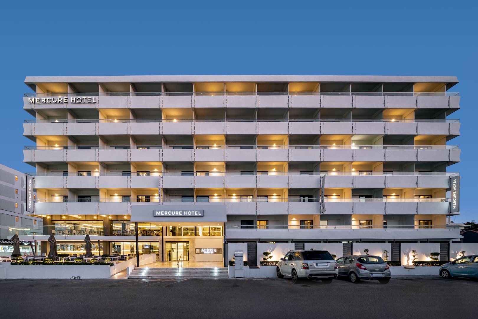 Mercure Rhodes Alexia Hotel & Spa ēkas fasāde vakarā ar izgaismotiem balkoniem un ieeju Rodas pilsētā, Grieķijā