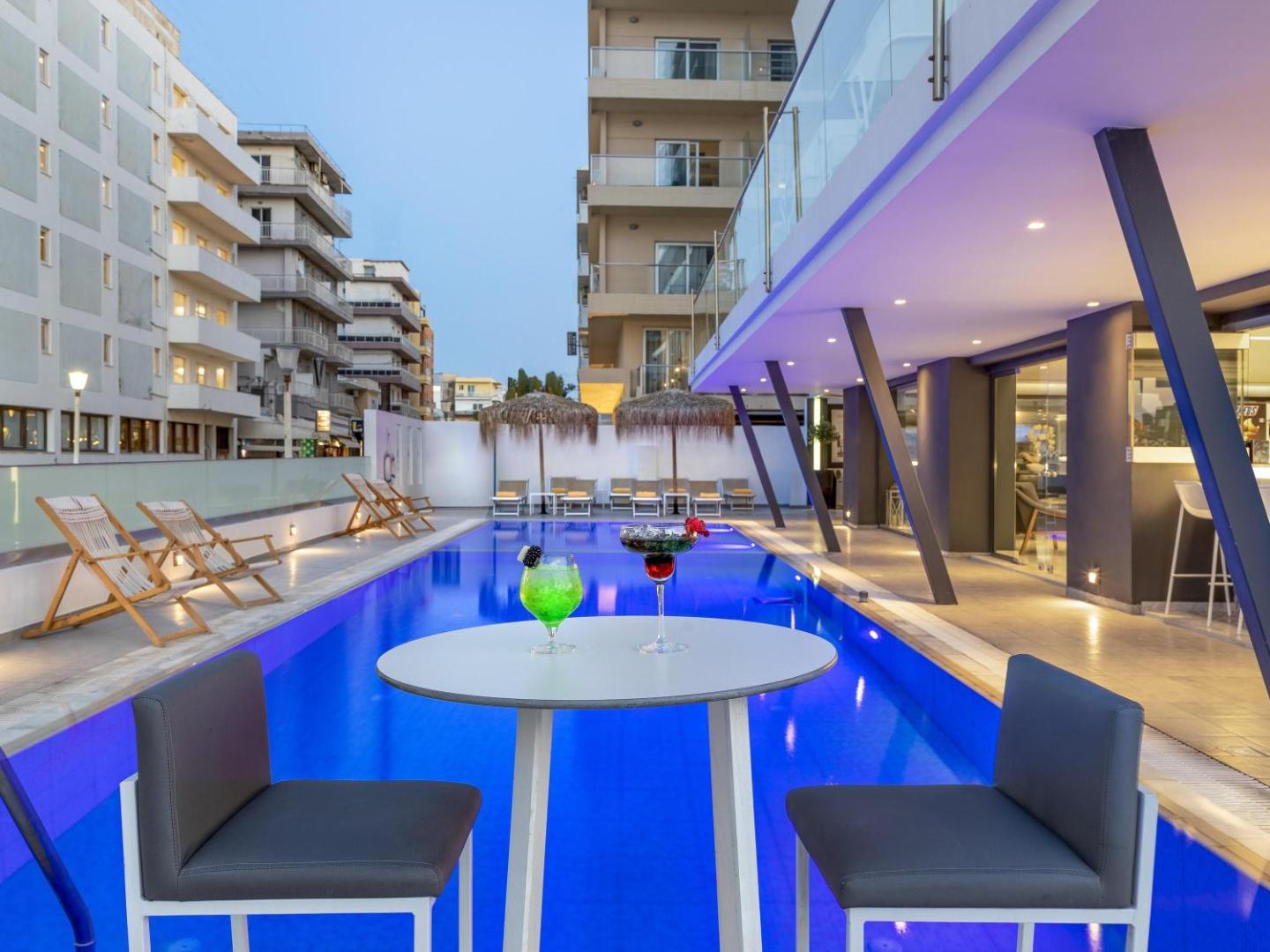 Mercure Rhodes Alexia Hotel & Spa āra baseins vakarā ar kokteiļiem priekšplānā un pilsētas arhitektūru fonā Rodas pilsētā.