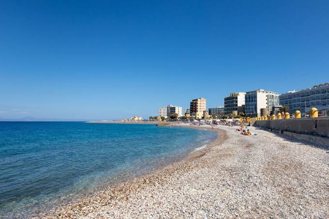 Mercure Rhodes Alexia Hotel & Spa tuvumā esošā oļu pludmale Rodas pilsētā ar zilu jūru un pilsētas ēkām fonā.