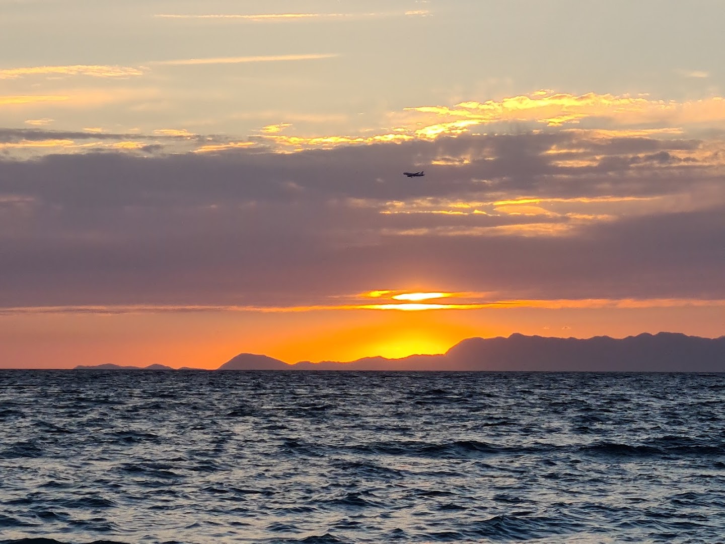 Saulriets virs Egejas jūras pie Sirene Beach Hotel Rodā — oranžas debesis, kalnu silueti tālumā un lidmašīna gaisā