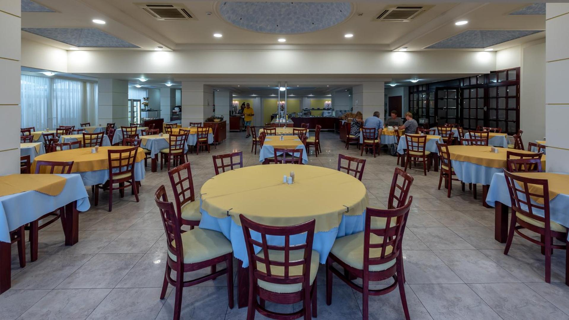 Sirene Beach Hotel galvenais restorāns ar klātiem galdiem, dzelteniem galdautiem un bufetes zonu fonā