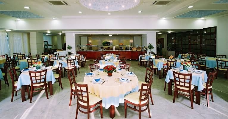 Sirene Beach Hotel galvenais restorāns Rodas salā — plaša telpa ar klātiem galdiem, koka krēsliem un bufetes zonu fonā