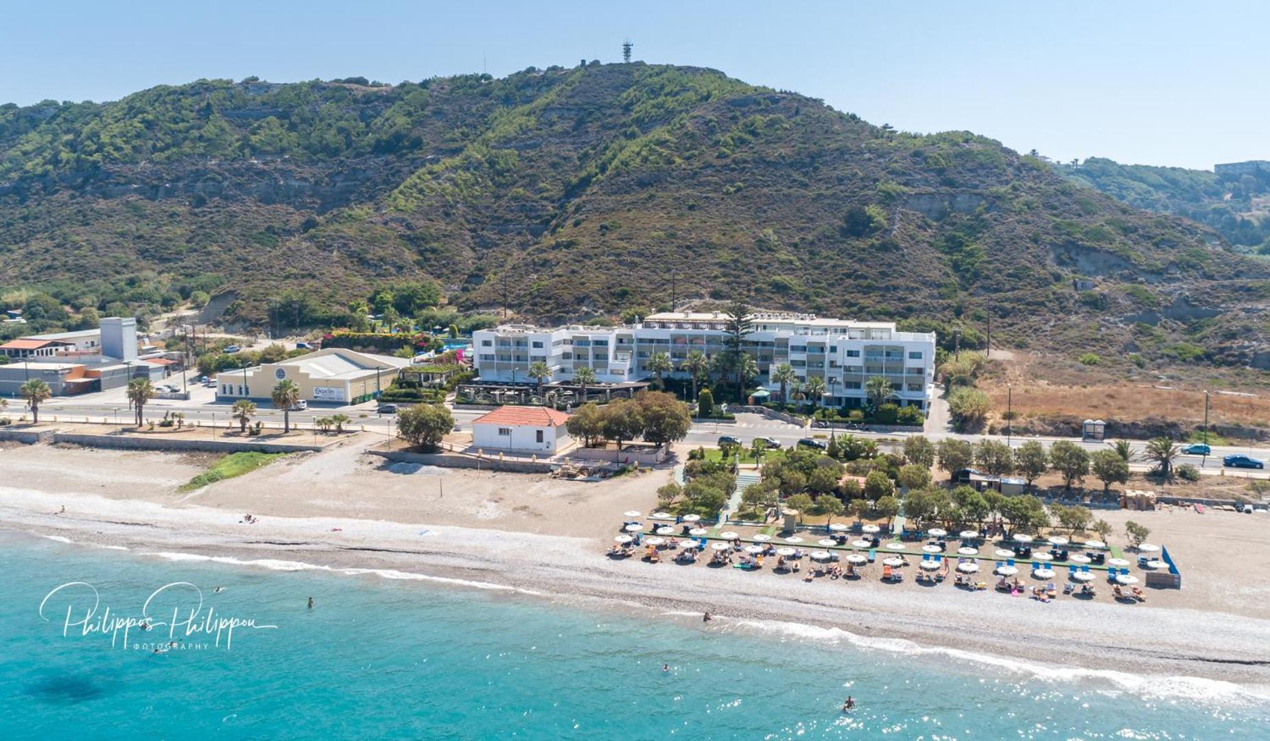 Sirene Beach Hotel panorāmas skats no jūras puses uz viesnīcas kompleksu, labiekārtotu pludmali ar saulessargiem un kalnu fonā