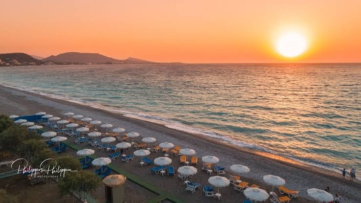 Sirene Beach Hotel panorāmas skats uz pludmali un saulrietu Rodā — rindu rindām izkārtoti saulessargi un oļu krasts
