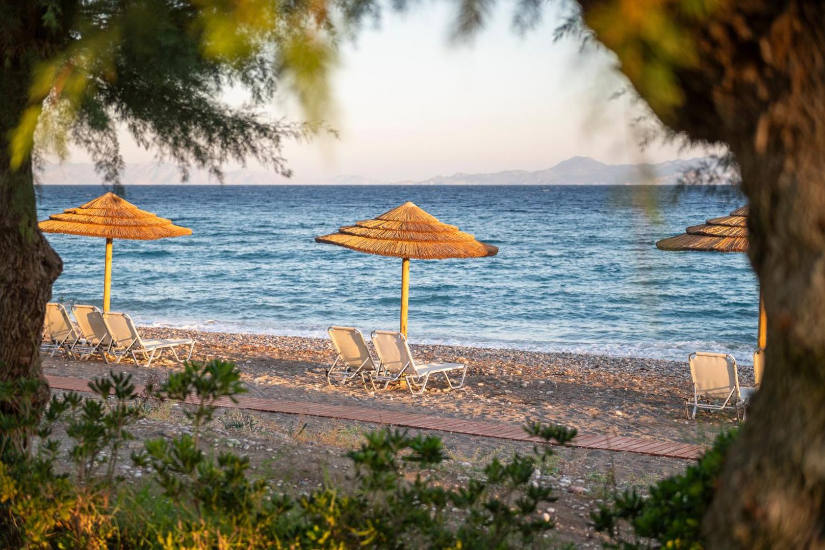 Skats caur koku zariem uz D' Andrea Mare Hotel pludmali ar salmu saulessargiem un zviļņiem Rodas salā