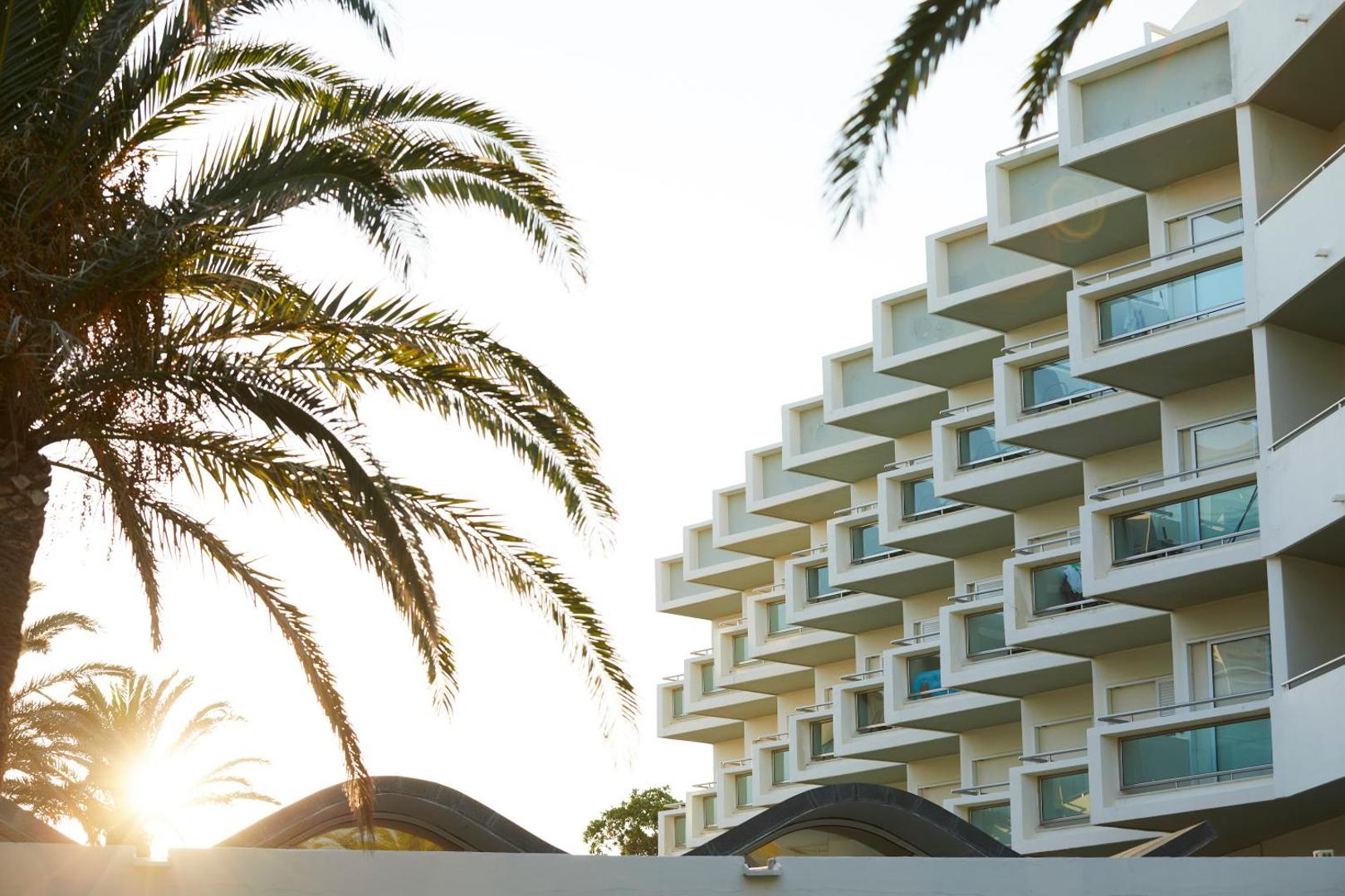 Apollo Beach viesnīcas modernā fasāde ar daudzlīmeņu balkoniem saulrieta gaismā — priekšplānā palmu lapas uz gaišo debesu fona.