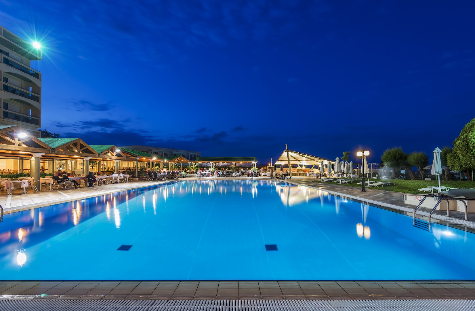 Apollo Beach viesnīcas lielais āra baseins vakarā Rodas salā — izgaismota terase ar restorāna galdiņiem un vakariņojošiem viesiem