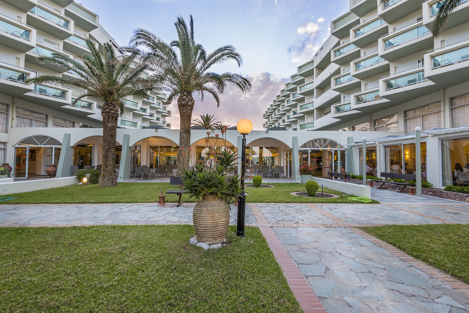 Apollo Beach viesnīcas centrālais laukums vakarā ar apgaismotām terasēm, palmām un dekoratīviem augiem — skats uz restorāna zonu un korpusiem.