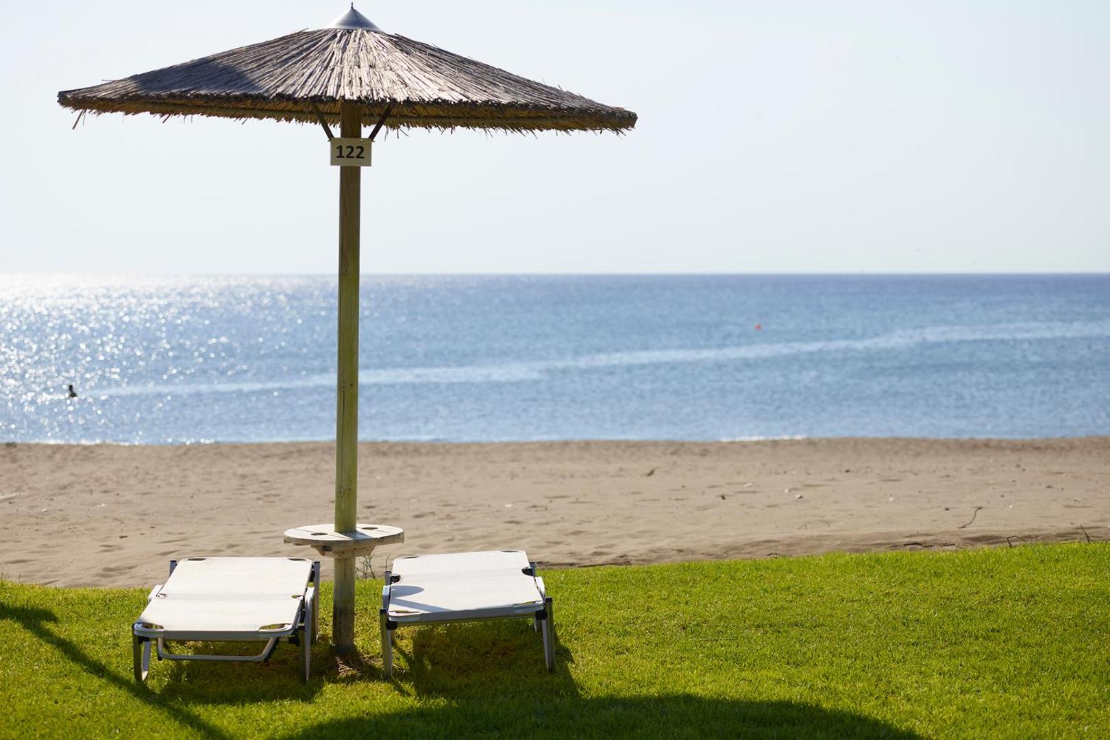 Apollo Beach viesnīcas sauļošanās krēsli un saulessargs uz zālāja pie pludmales Faliraki, Rodas salā ar skatu uz jūru