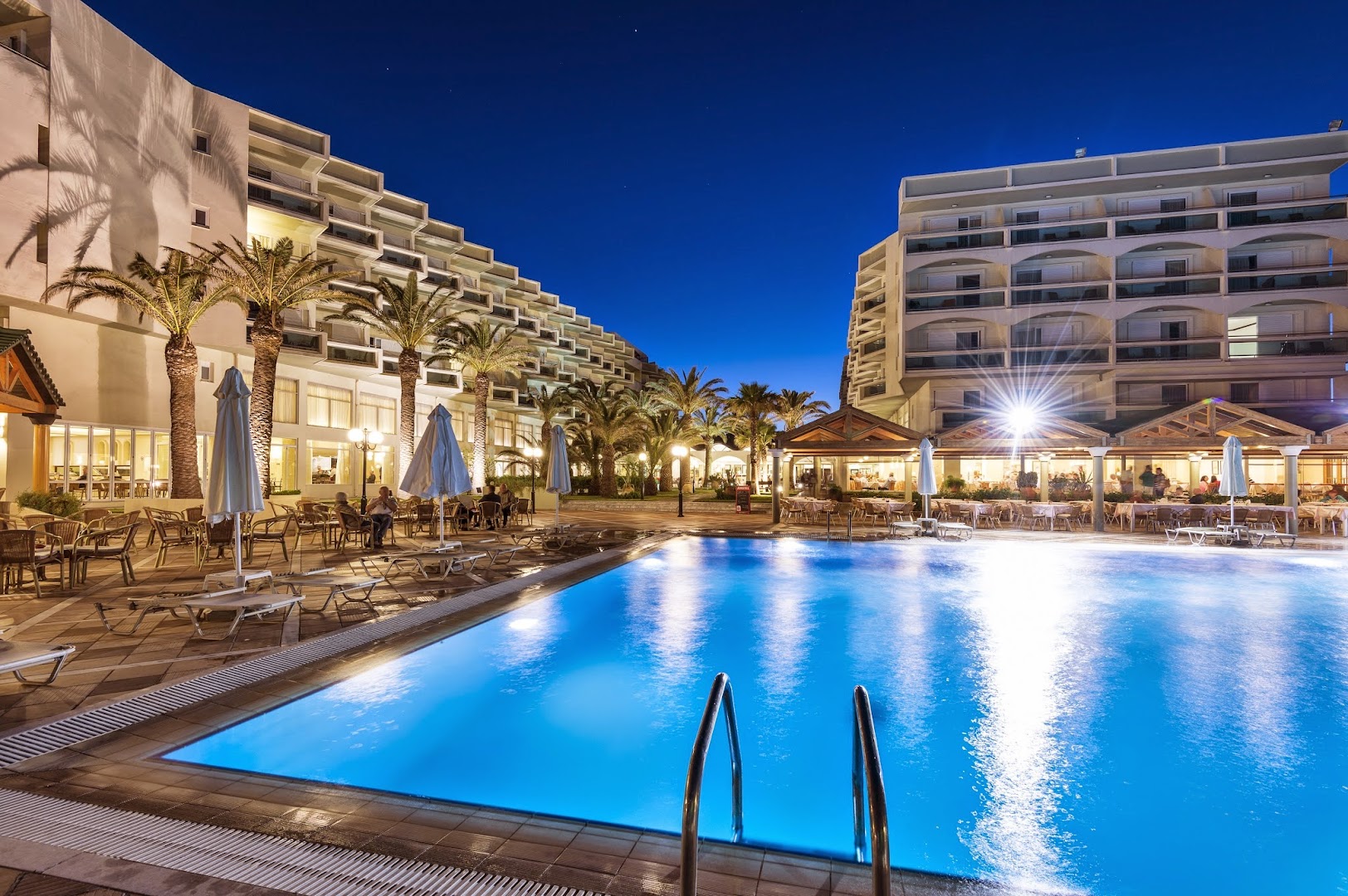 Apollo Beach kūrorta baseins un restorāna terase naktī ar izgaismotām viesnīcas ēkām un palmām Faliraki