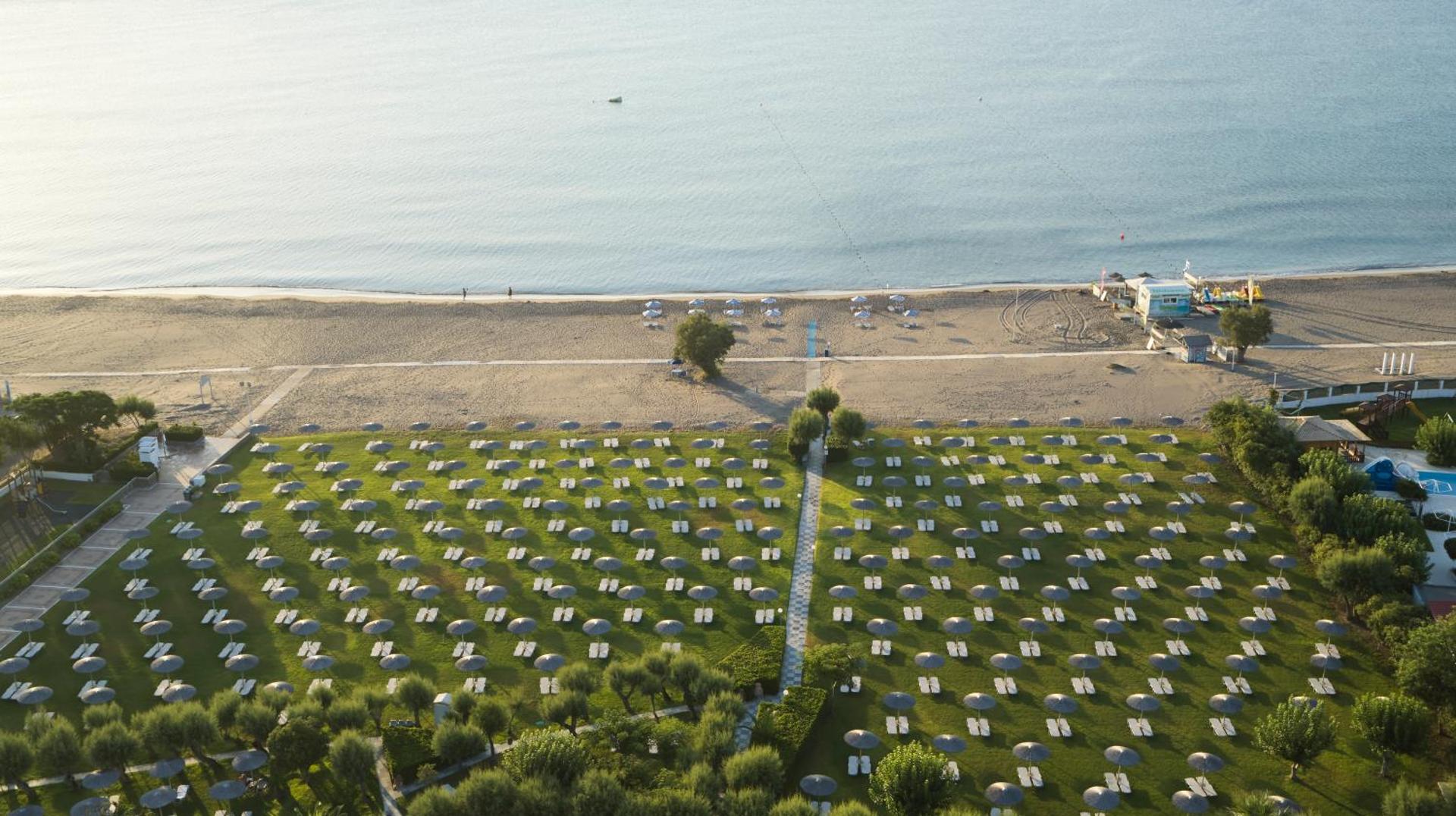 Apollo Beach viesnīcas dārzs ar daudziem saulessargiem zālienā un skatu uz jūru — aerofoto uzņēmums rīta gaismā
