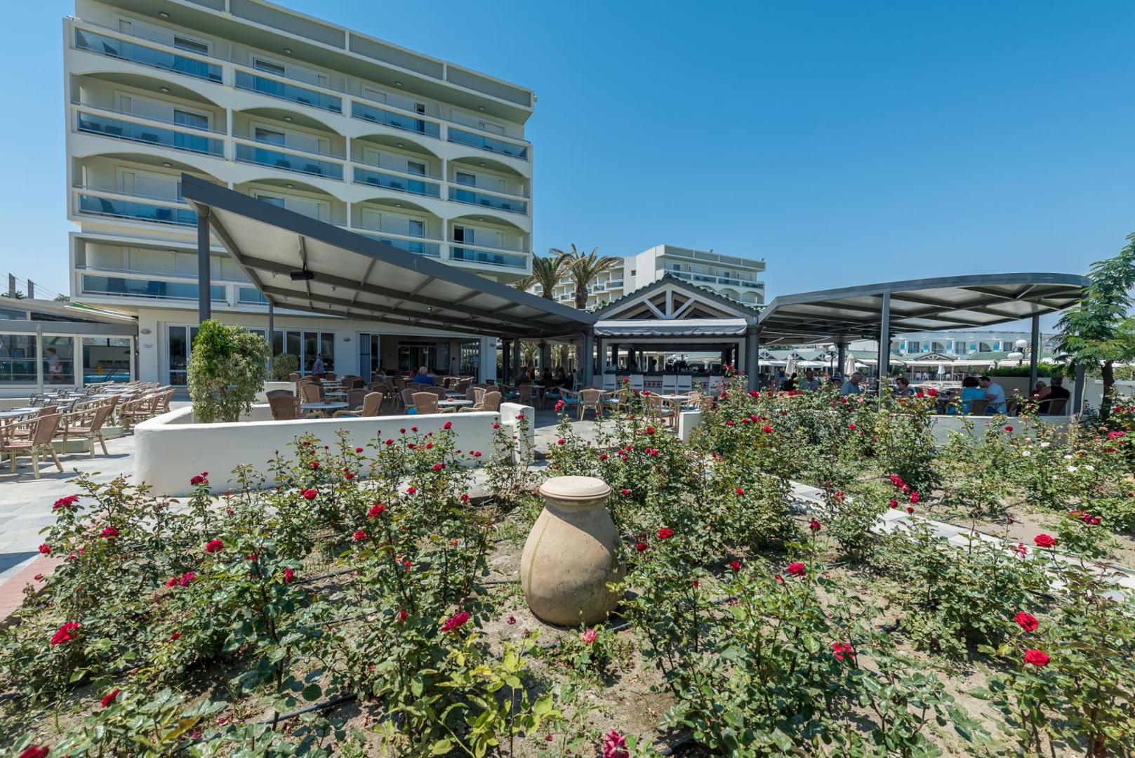 Apollo Beach viesnīcas āra terase Rodas salā ar ziedošām rozēm priekšplānā un galdiņiem zem nojumes pie galvenās ēkas