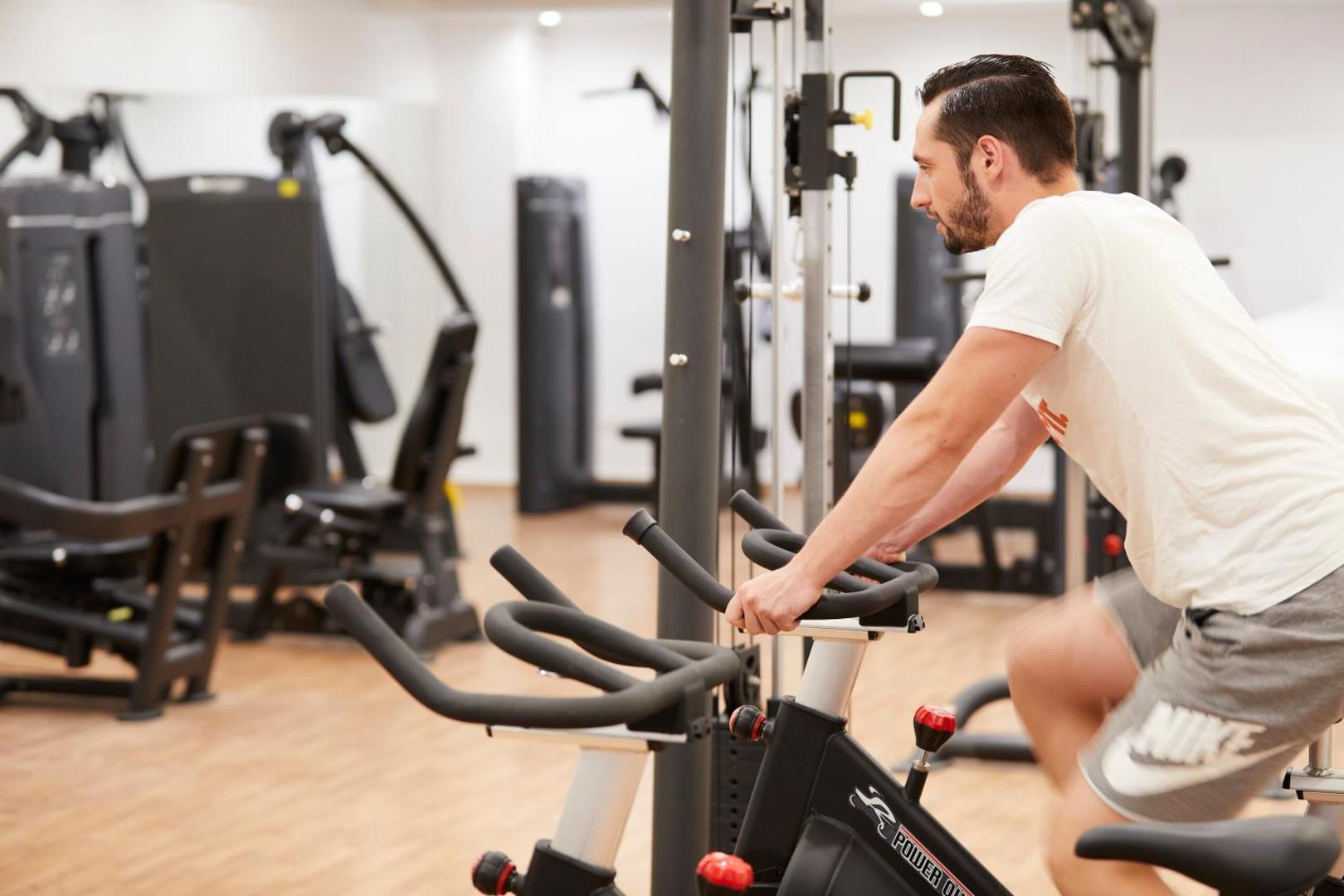 Viesis sporto Apollo Beach viesnīcas fitnesa centrā uz velotrenažiera, fonā redzams mūsdienīgs sporta zāles aprīkojums